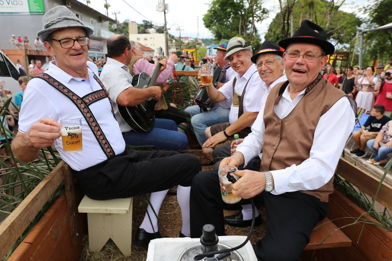Senhores de chapéu e colete típico com canecas de chope na carroça do desfile